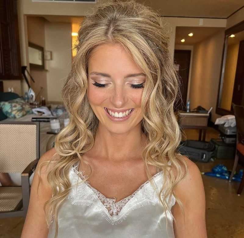 Bride in a satin dress smiling, preparing for her wedding in a hotel room, with curled blonde hair.